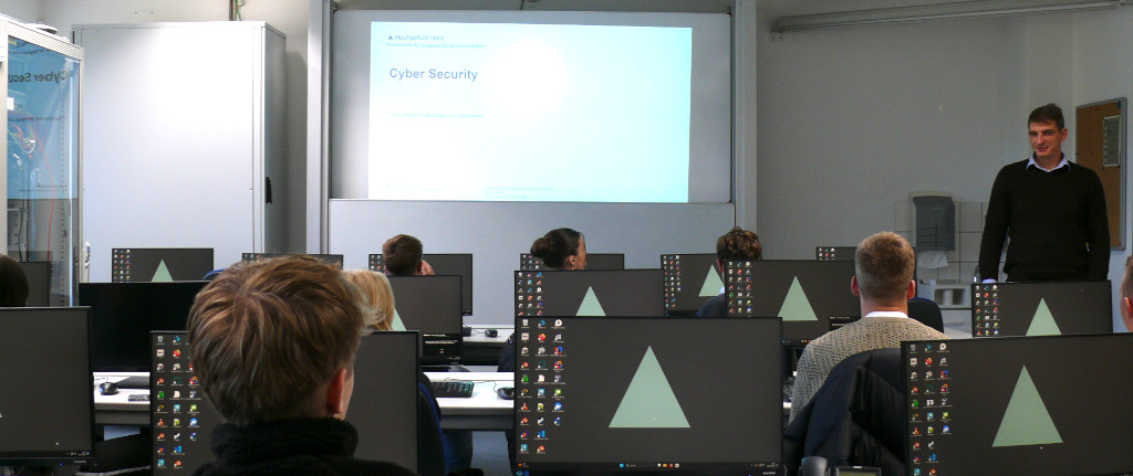 Schüller sitzen an Tischen mit Rechnern, Professor Rempel erklärt am Whiteboard Cyber Security