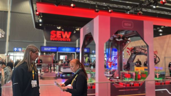 Frau sortiert Flyer in ihrer Hand, gegenüber steht ein Mann, im Hintergrund große Messestand mit Robotertechnik